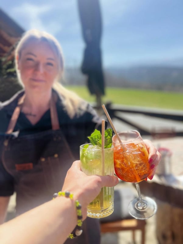 Anstoßen mit Mojito und Spritz auf der sonnigen Terrasse, im Hintergrund das Personal und grüne Wiesen