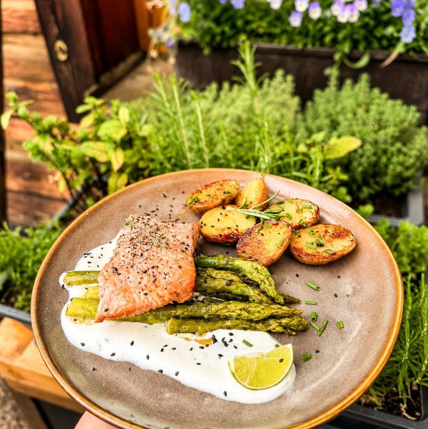 Gebratener Lachs mit Spargel, Kartoffeln und Joghurtsoße auf einem Teller zwischen Kräutern auf der Terrasse