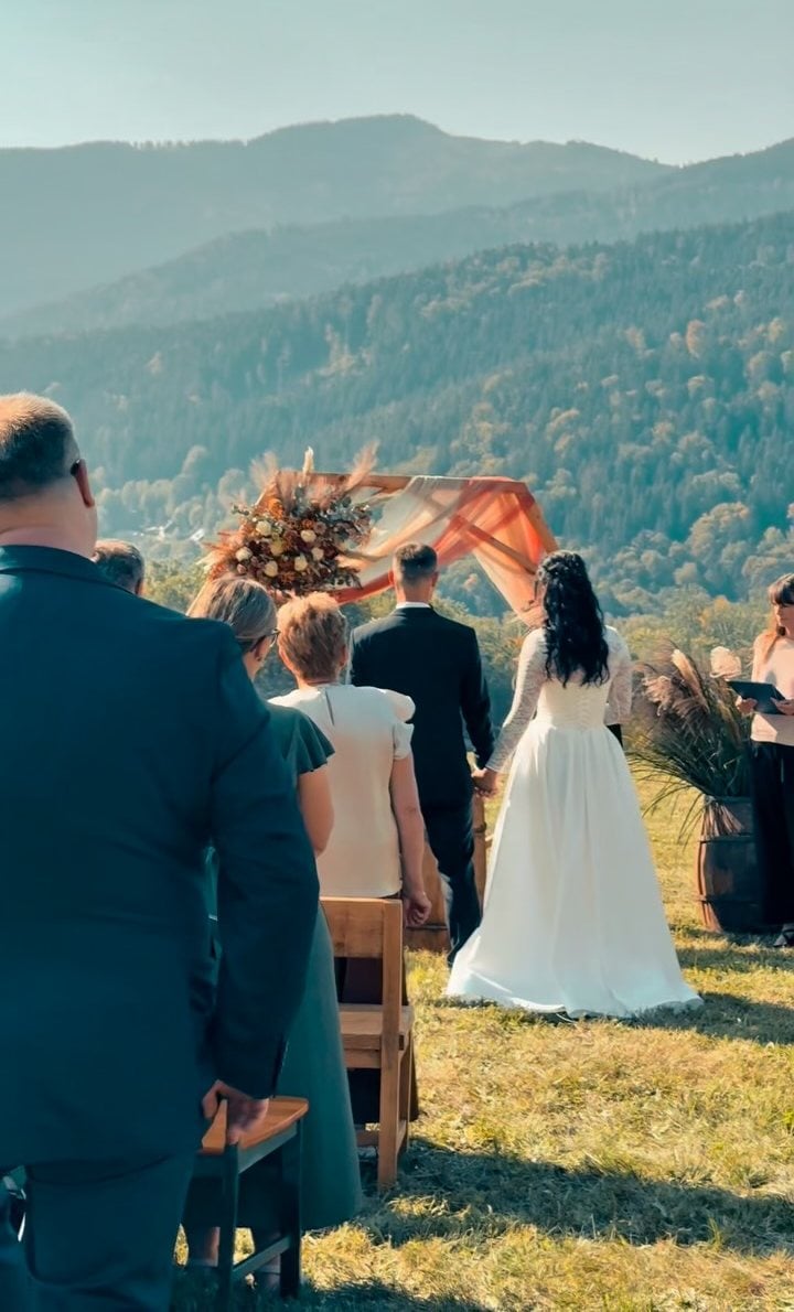 Svatební obřad na louce, nevěsta a ženich u květinové brány, za nimi zalesněné hřebeny Šumavy