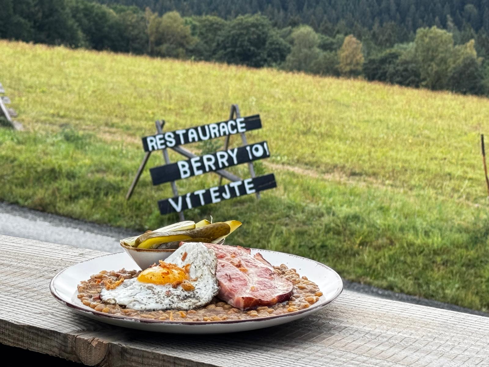 Talíř s čočkou, uzeným a volským okem na zábradlí terasy, za ním cedule „Restaurace Berry vítejte“